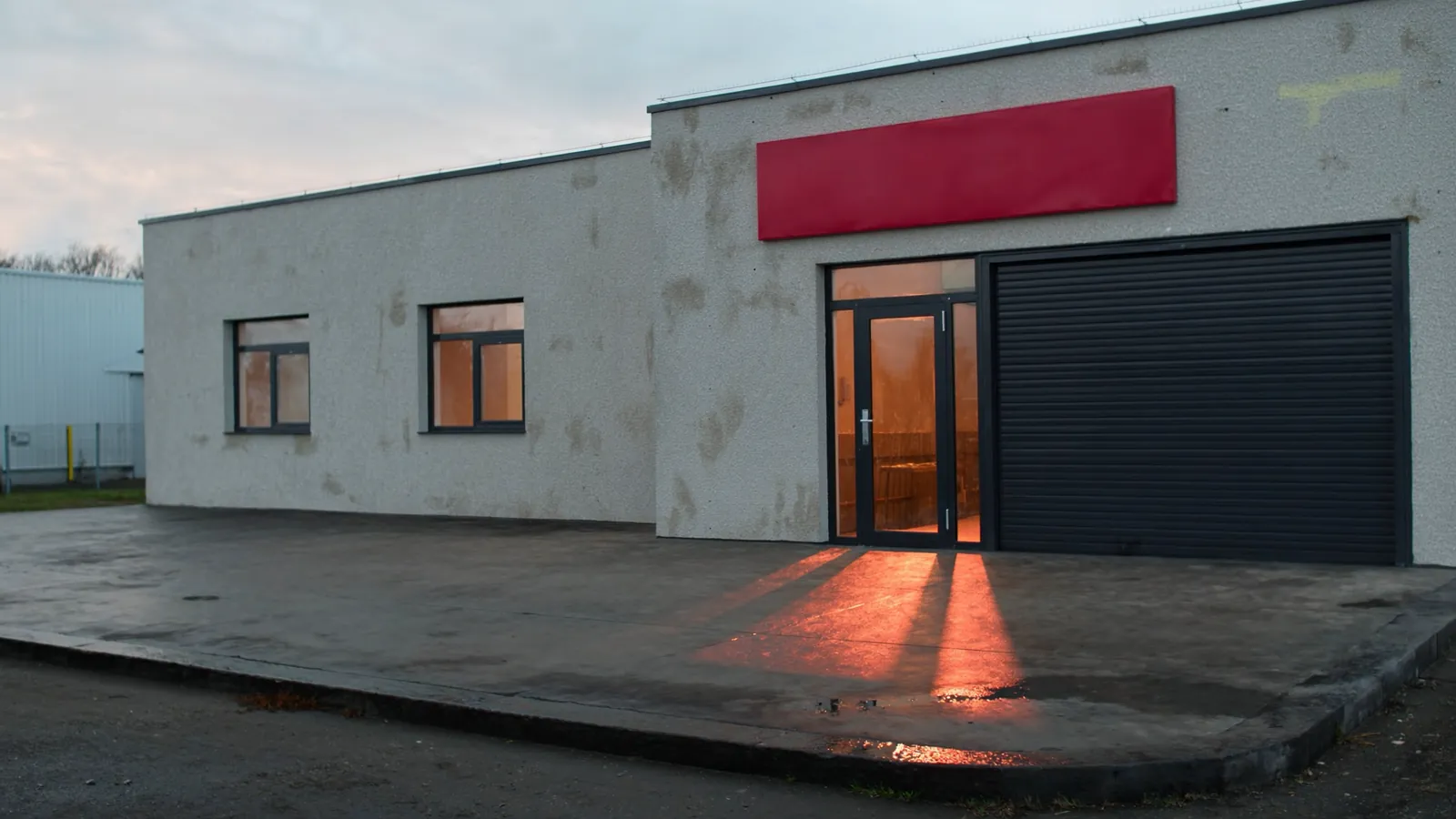 The garage premises at dusk: light grey concrete block walls, dark metal frames and an entrance lit with warm light onto wet tarmac